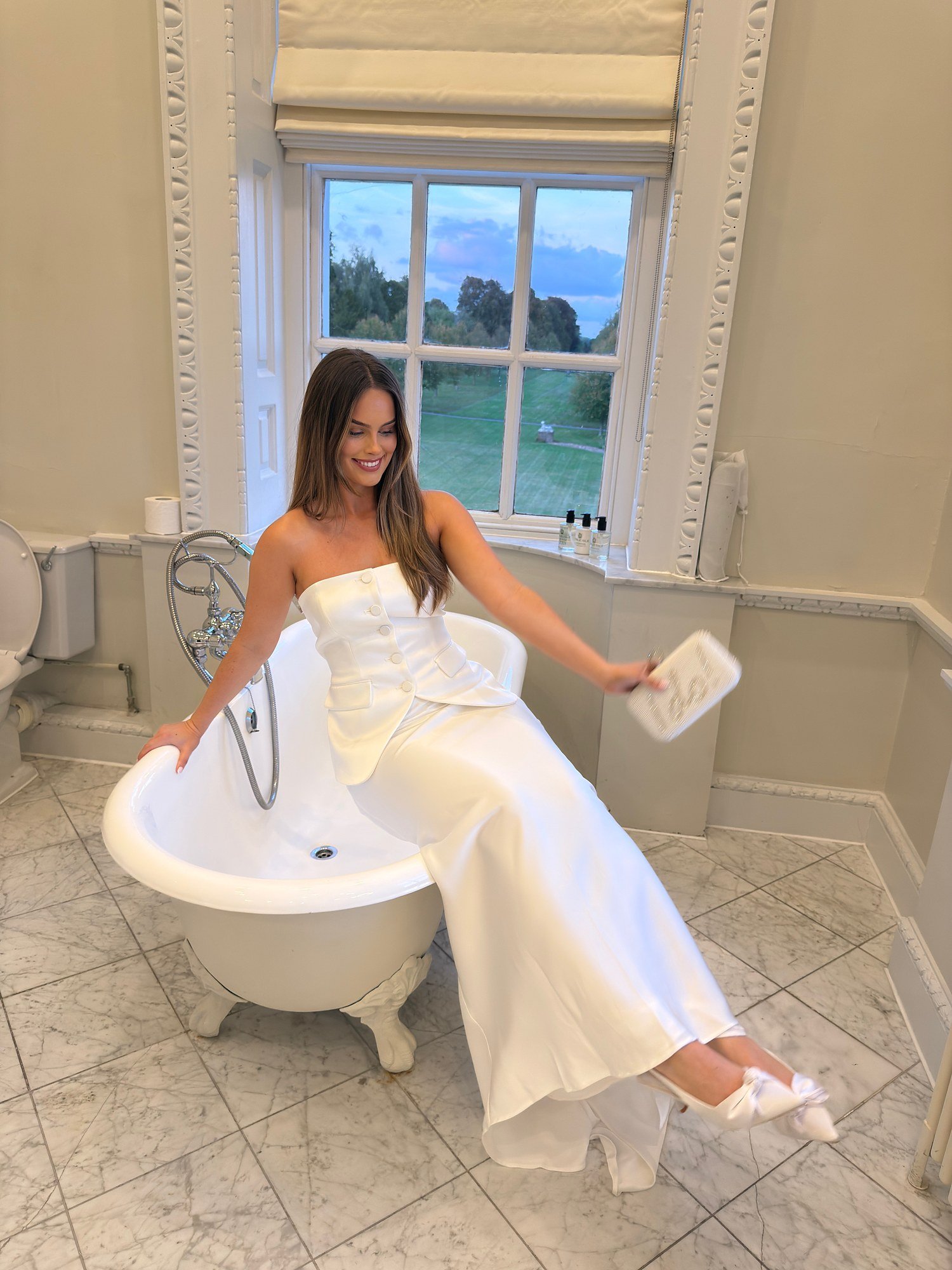 Bride in a strapless satin gown seated by a window with parkland behind