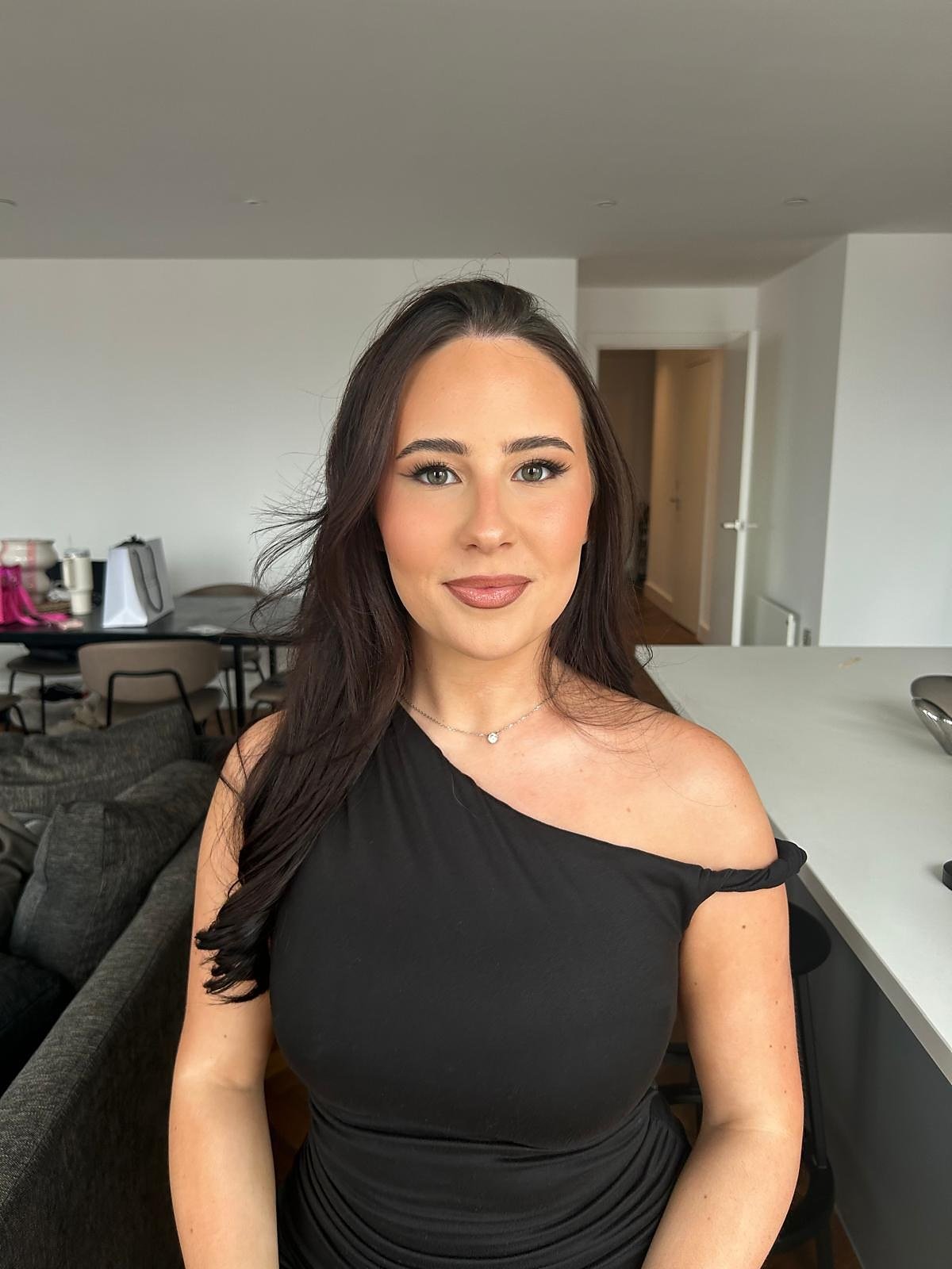 Occasion makeup look: client in a black one-shoulder dress with long dark waves, soft polished makeup and a nude lip