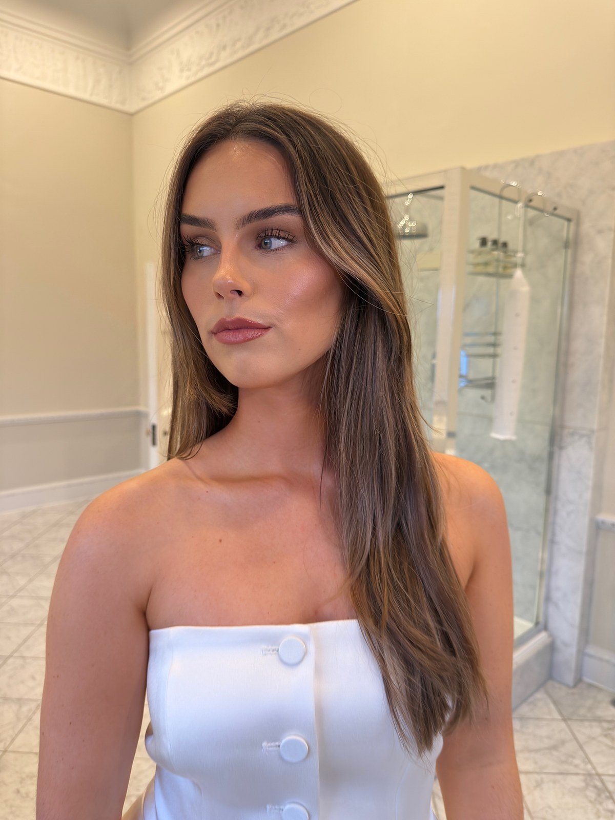 Bridal makeup look: client in a white strapless top with long brunette hair, soft glowing skin and a nude lip, in a marble bathroom
